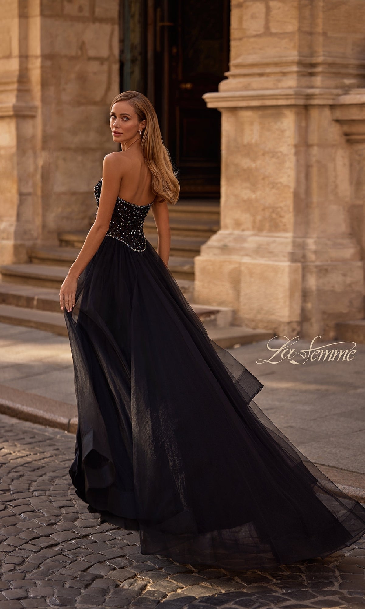 A woman in the La Femme 32856 Strapless Long Black Prom Ball Gown with a beaded bodice stands on a cobblestone street before a stone building, elegantly glancing over her shoulder.